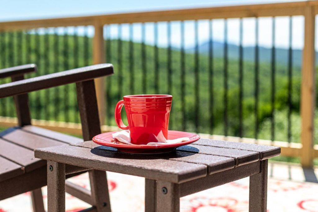A wooden table with a red cup and saucer on it, set on a balcony with a view of green hills.