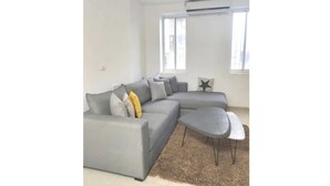 Un salon moderne avec un canapé d’angle gris, une petite table basse et une grande fenêtre qui laisse entrer la lumière naturelle.