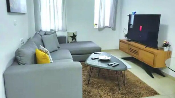 Un salon avec un canapé gris, une table basse, un téléviseur posé sur un meuble en bois et une fenêtre avec des rideaux.