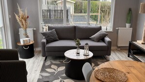 A modern living room with a grey sofa, a round wooden table, and a black coffee table. There are two black chairs, a black storage unit, and a potted plant.
