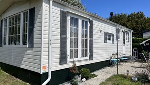 Il s’agit d’une maison mobile de plain-pied, avec une façade verte, un revêtement blanc et des volets noirs. Elle est dotée d’un petit porche à l’avant, avec une chaise et une table, ainsi que d’une allée en gravier.