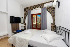 A bedroom with a bed, a flat-screen TV, a stone wall, and a door leading to a balcony. - One bedroom apartment with free car rental (Heated Pool) (Περίσσα)