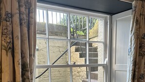 Quarto com janela em sacada, um banquinho e vista para uma escadaria de pedra externa.