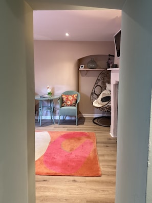 A cozy living room with a wooden floor, a colorful rug, a wicker chair, a small table with a decorative item, and a wall-mounted shelf with a vase and a glass.
