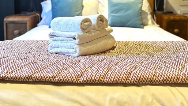 Uma cama bem arrumada com toalhas dobradas, um edredom estampado e uma parede com um letreiro de neon que diz 'amour'.