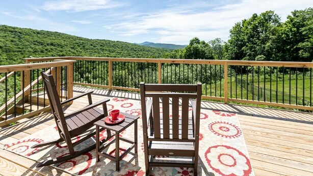 Une terrasse en bois avec une table et des chaises, un tapis à motifs et une vue magnifique sur des collines verdoyantes.