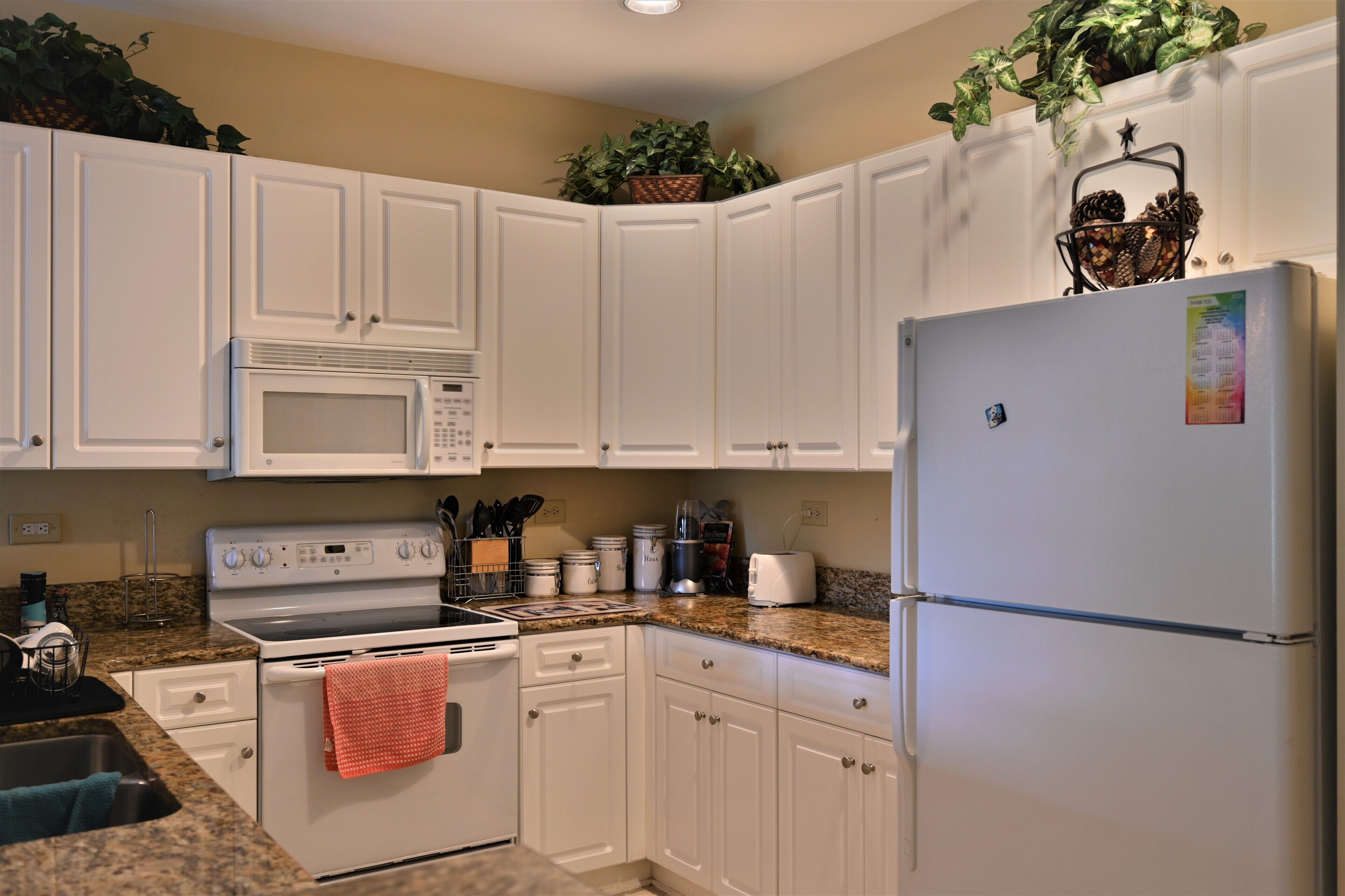 A kitchen with white cabinets, a microwave, stove, and refrigerator. There are plants on top of the cabinets and a basket with pinecones.