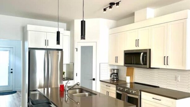A modern kitchen with stainless steel appliances, white cabinetry, and a central island with a sink. - The Northside Nest (Lethbridge)