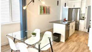 A modern kitchen with a central island, white cabinetry, stainless steel appliances, and a dining area with glass tables and chairs. - The Northside Nest (Lethbridge)