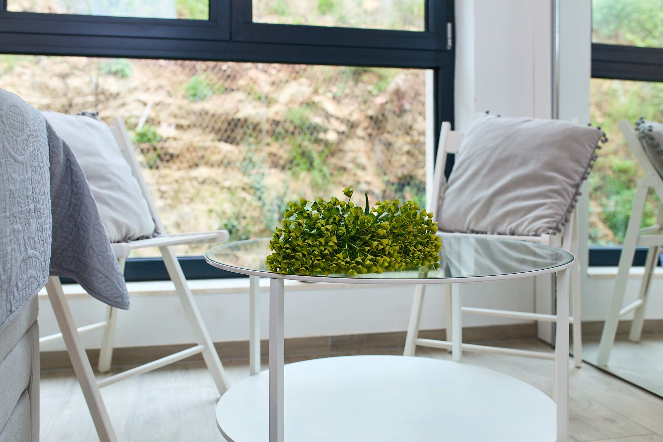A round glass table with a small plant, white chairs with cushions, and a view of a rocky outdoor area through a window.