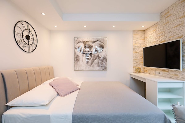 A bedroom with a bed, a clock, an elephant painting, a TV, and a stone-textured wall.
