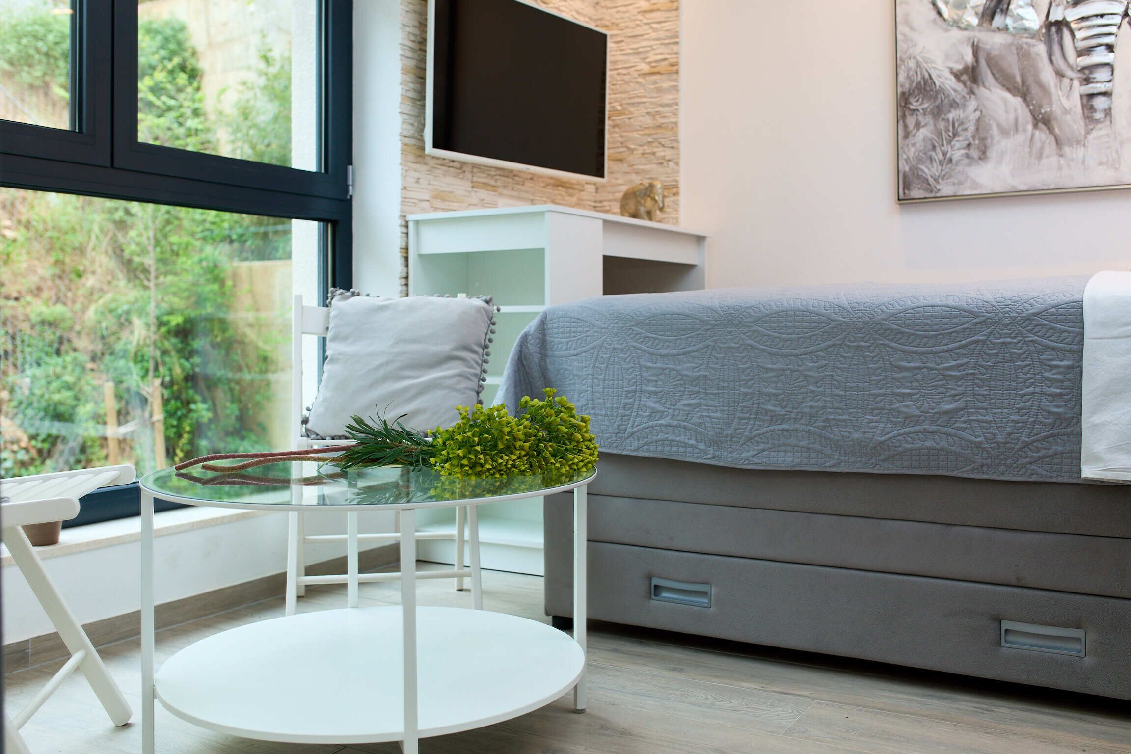 A modern bedroom with a bed, a TV mounted on the wall, a glass table with a plant, and a window with a view of greenery.