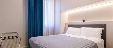 A modern hotel room with a neatly made bed, a bedside table, and a window with blue curtains.