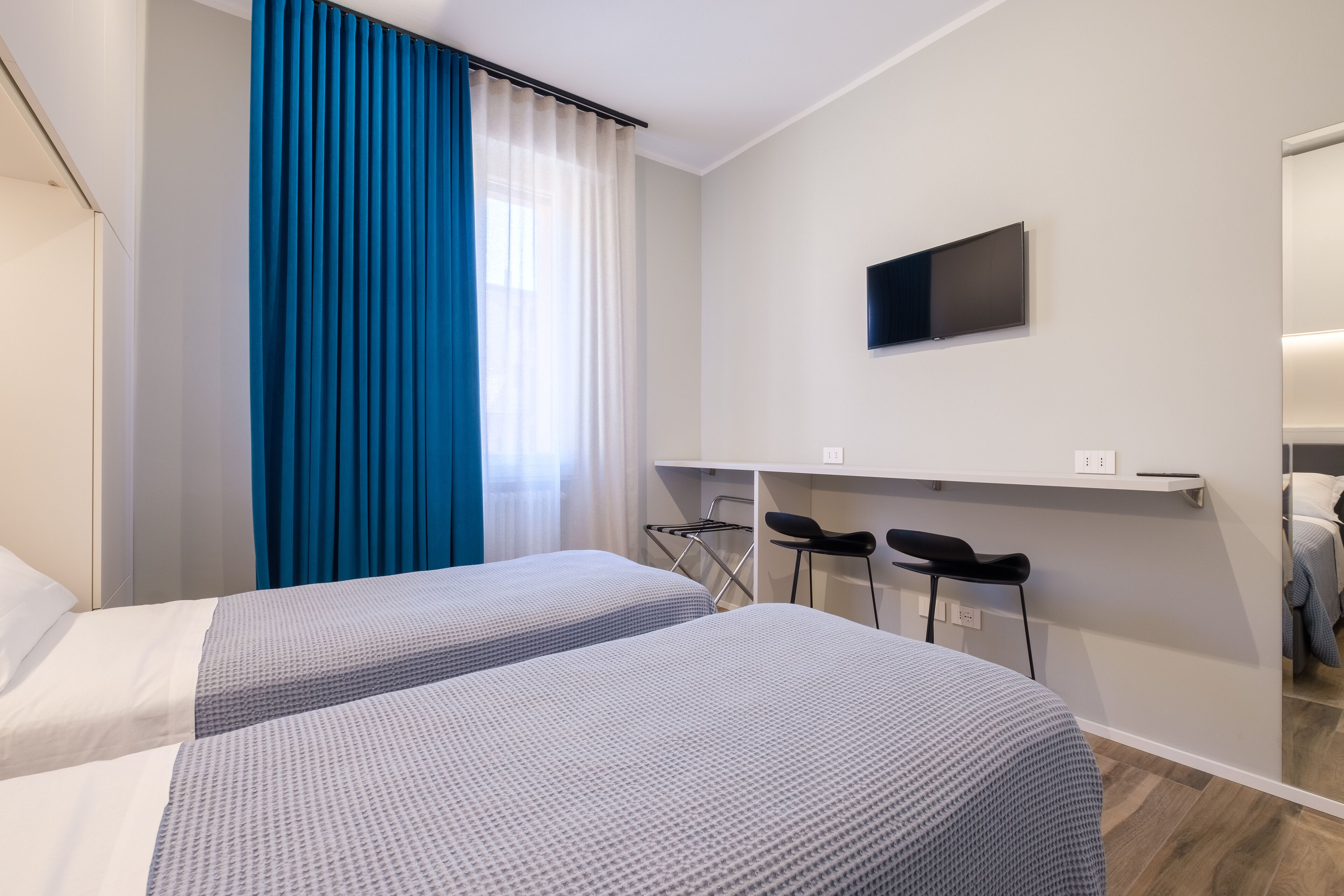 A hotel room with two beds, a TV mounted on the wall, a desk with chairs, and a window with curtains.