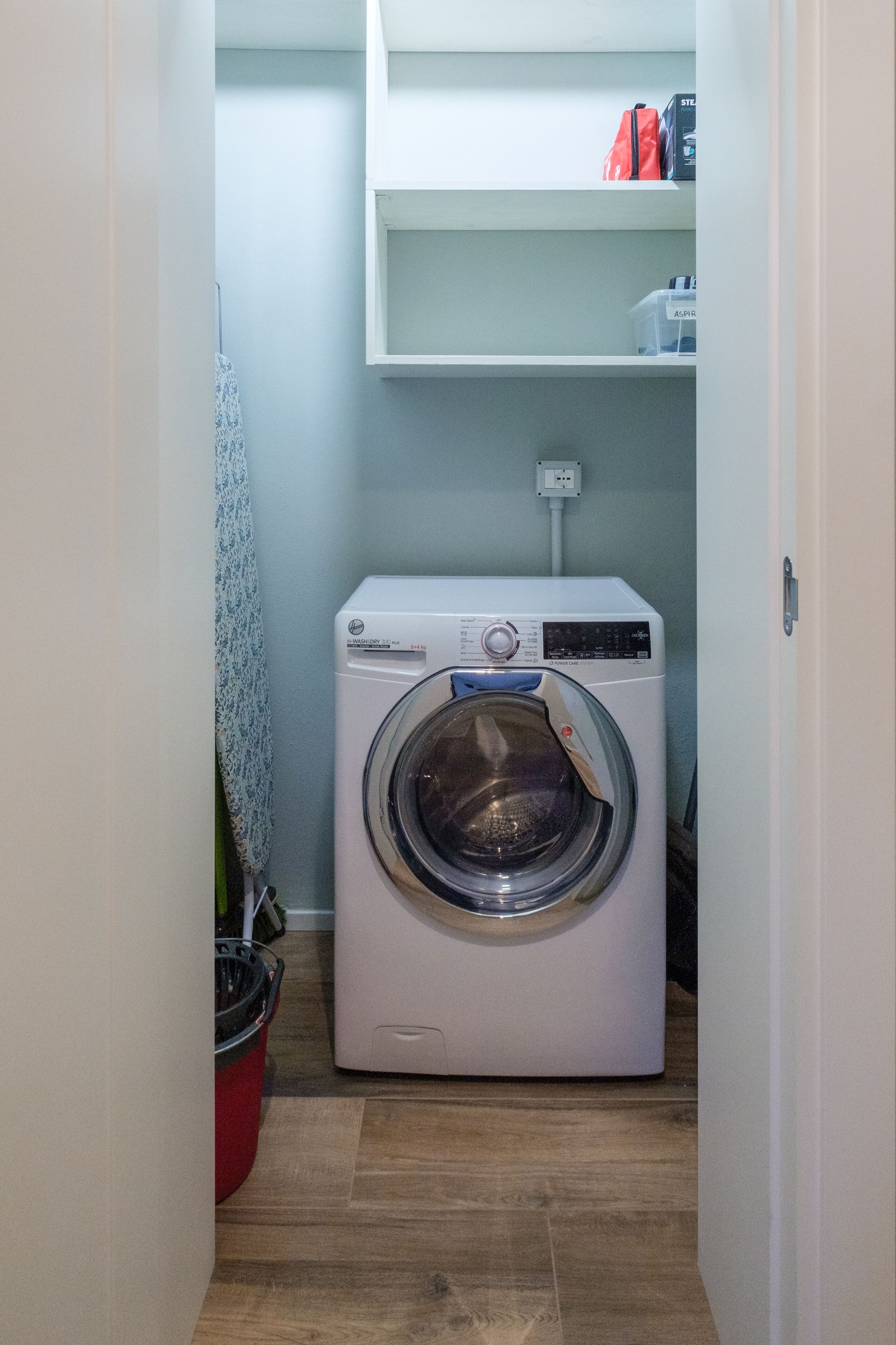 Une petite buanderie équipée d’un lave-linge à chargement frontal, d’une petite étagère au-dessus et d’une prise électrique sur le mur.