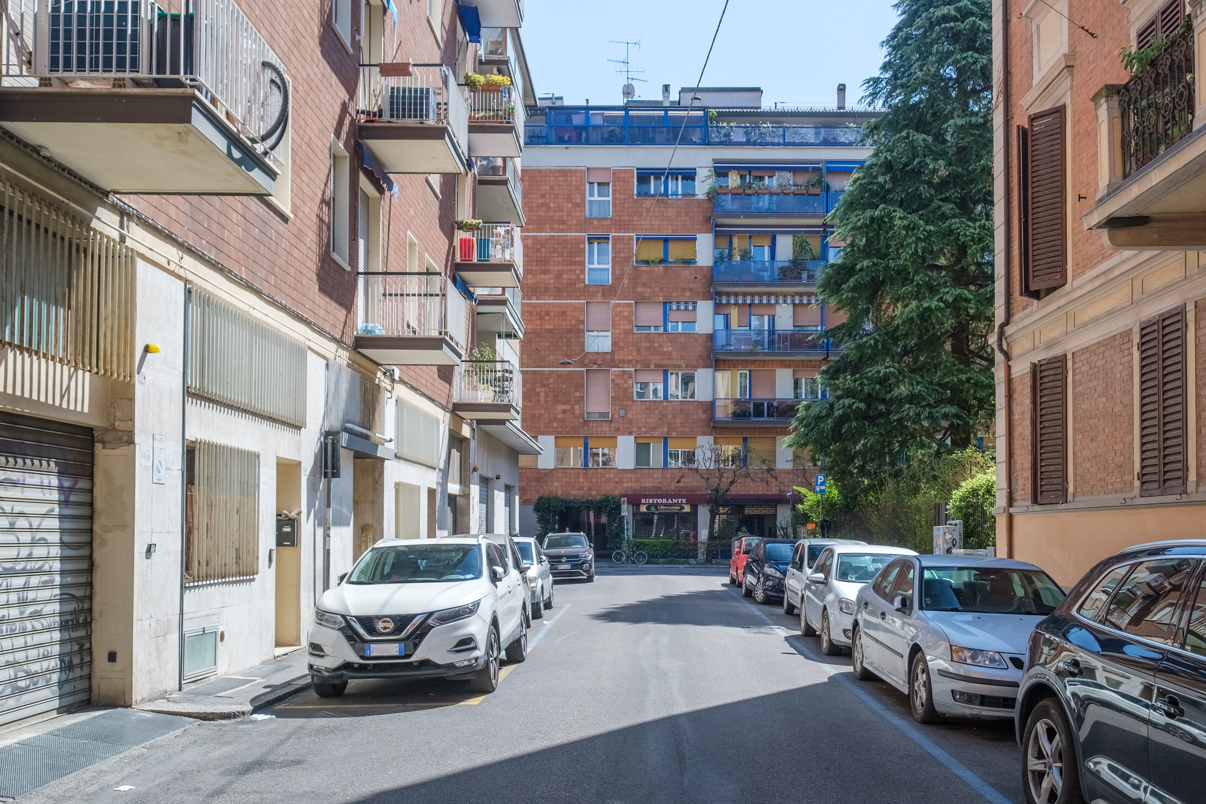 A narrow urban street lined with multi-story residential buildings, parked cars, and a restaurant.