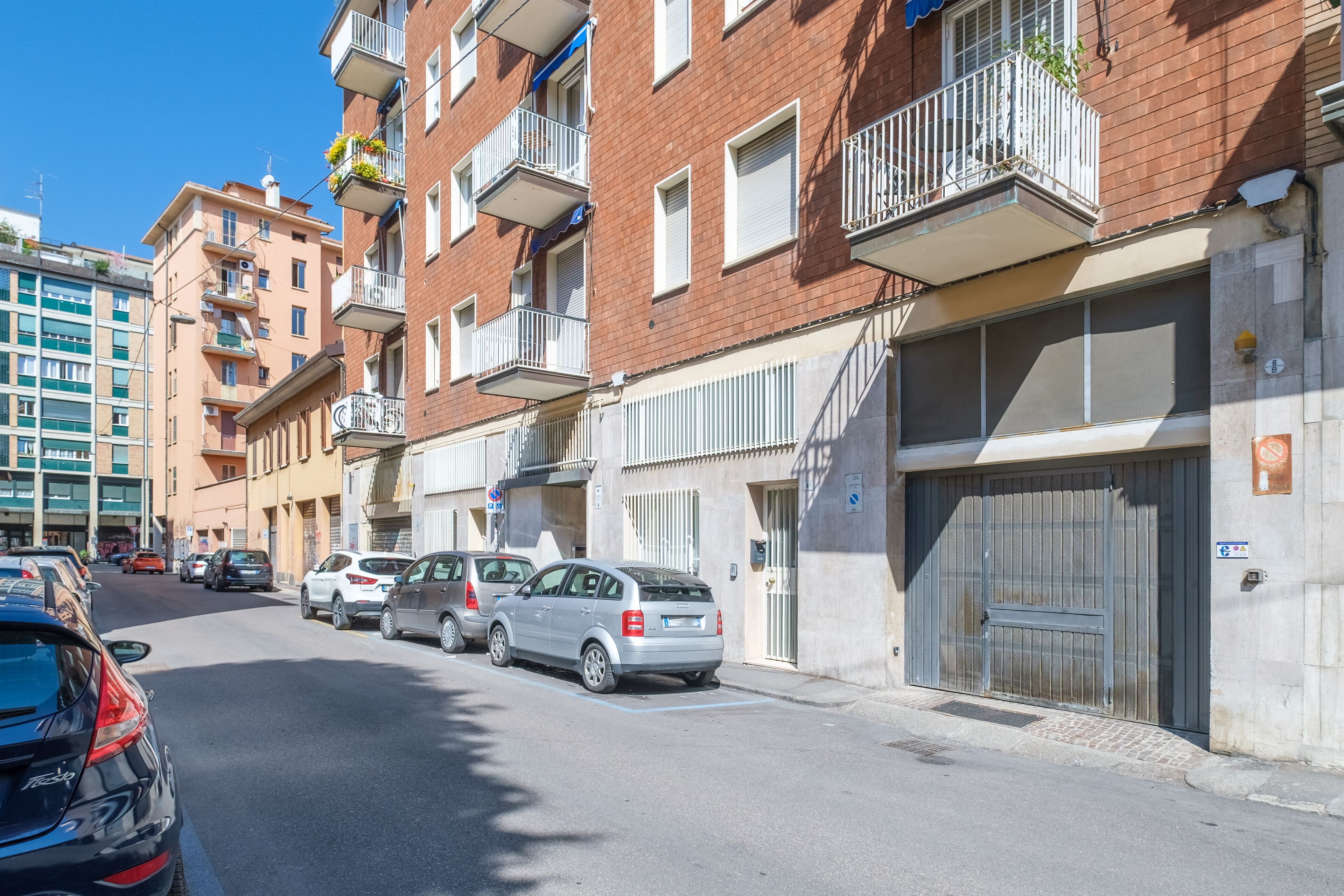 A row of multi-story residential buildings with balconies, parked cars, and a clear blue sky.