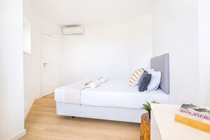 Un lit blanc avec des oreillers, un mur blanc, une porte et un appareil de climatisation.