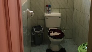 A small bathroom with a toilet, a trash bin, a green laundry basket, and a window with natural light. - B&B rent rooms  (Chaguanas)