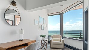 Une chambre moderne avec un bureau en bois, un miroir rond, une chaise, une petite table d’appoint avec une plante et une grande fenêtre donnant sur la mer.