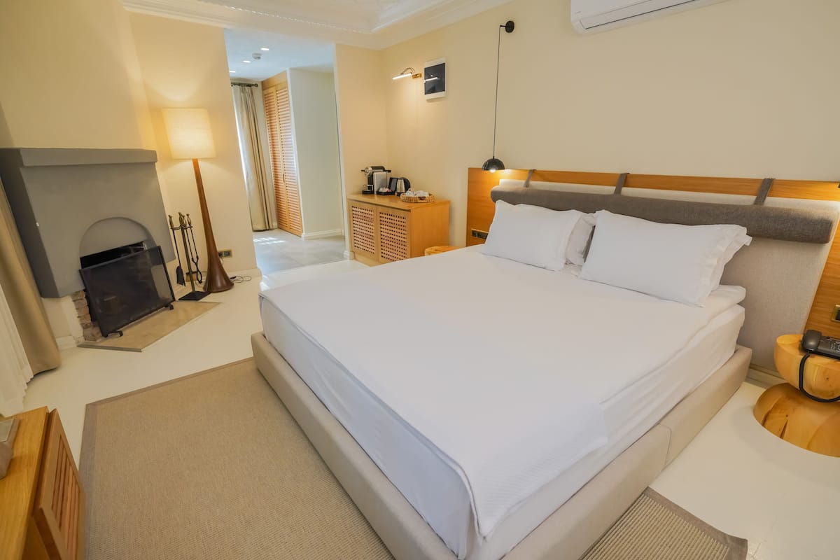 A hotel room with a large bed, a wooden headboard, a bedside table with a lamp, a small wooden cabinet, and a fireplace.