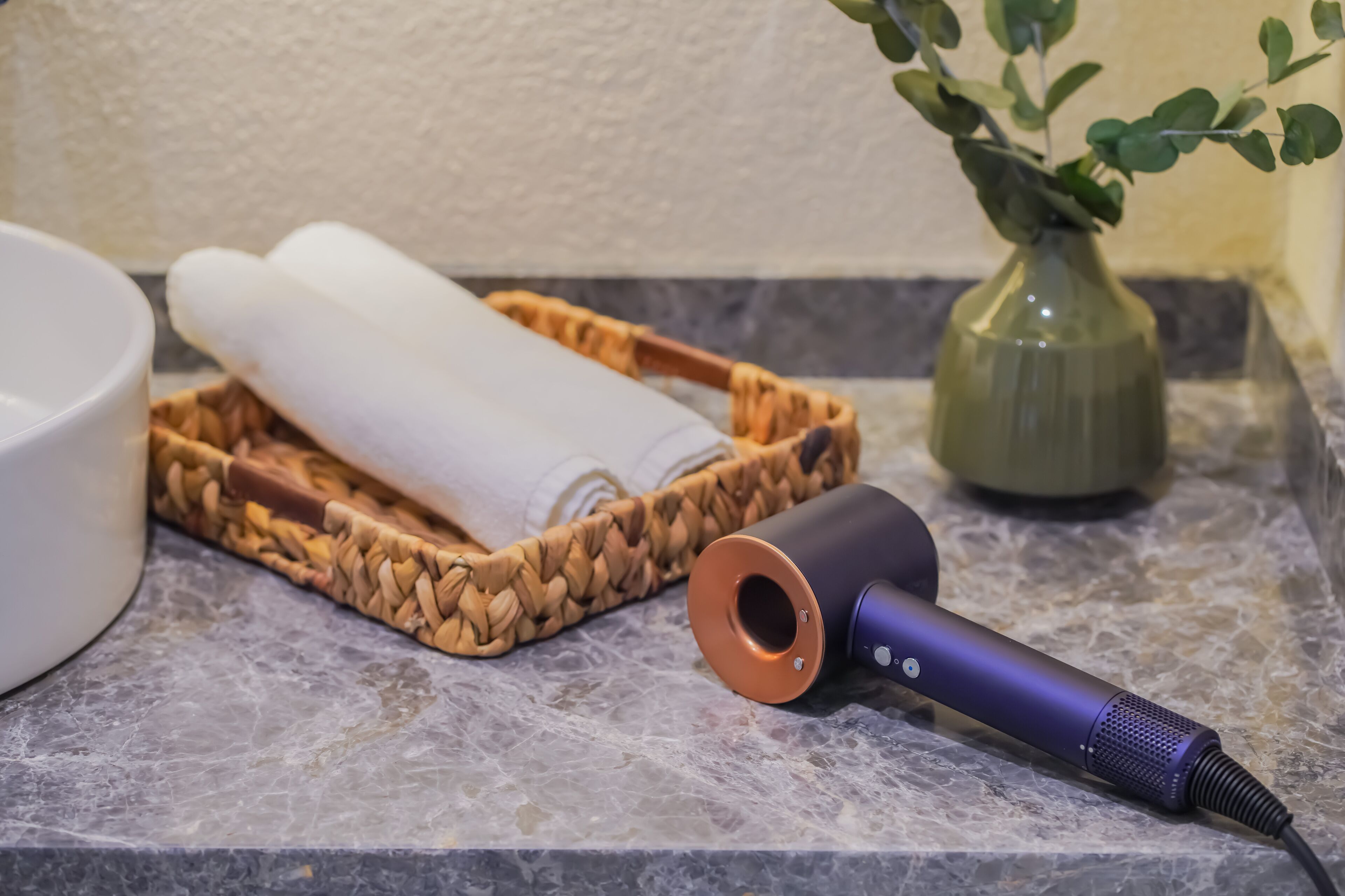 A bathroom with a marble countertop, a woven basket containing rolled towels, a green vase with a plant, and a purple handheld device.