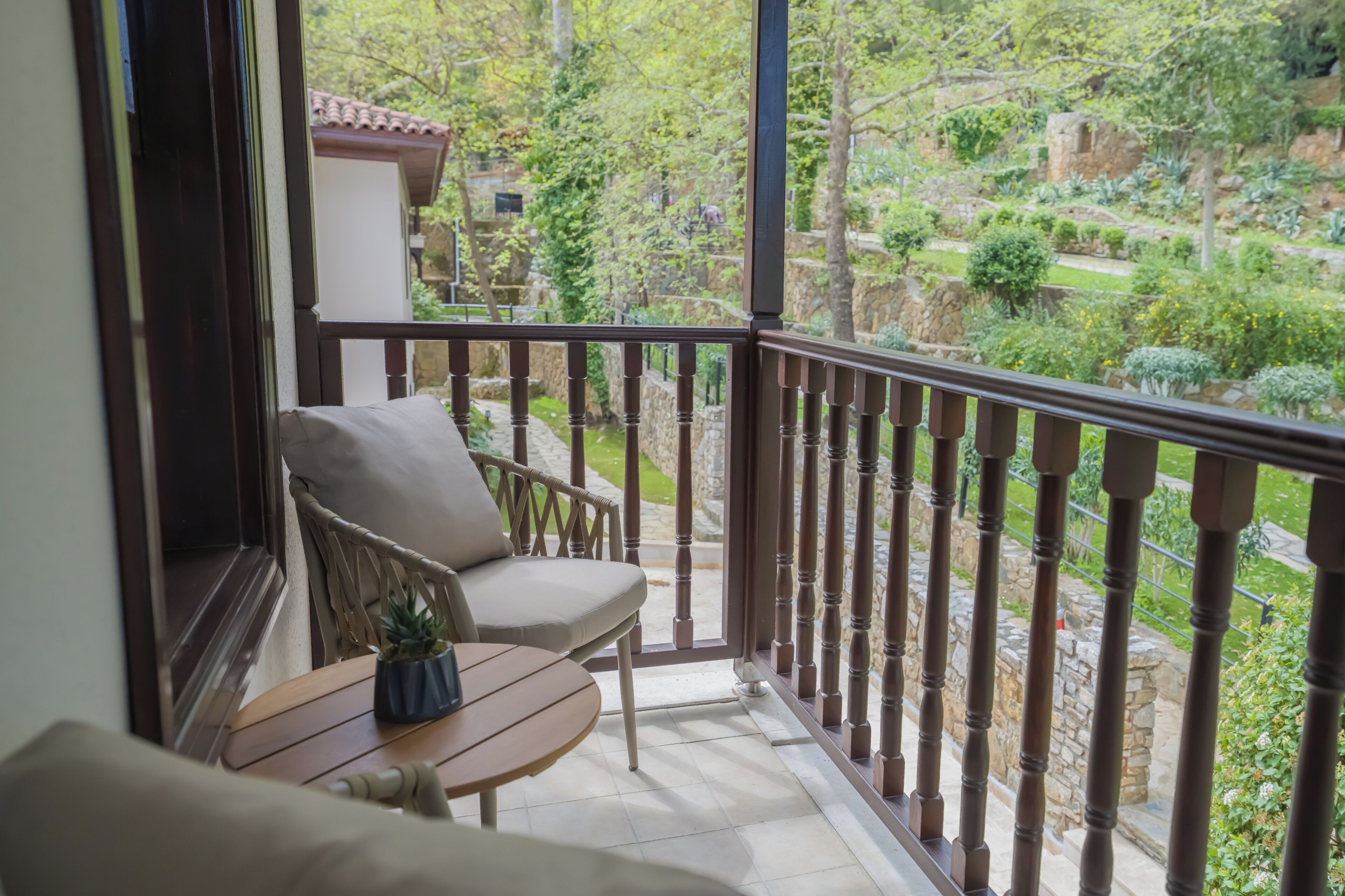 A balcony with a chair and table, a view of a garden with various plants and a stone wall.