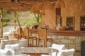A covered outdoor seating area with wicker chairs and tables, a stone wall, and a bar with bottles.
