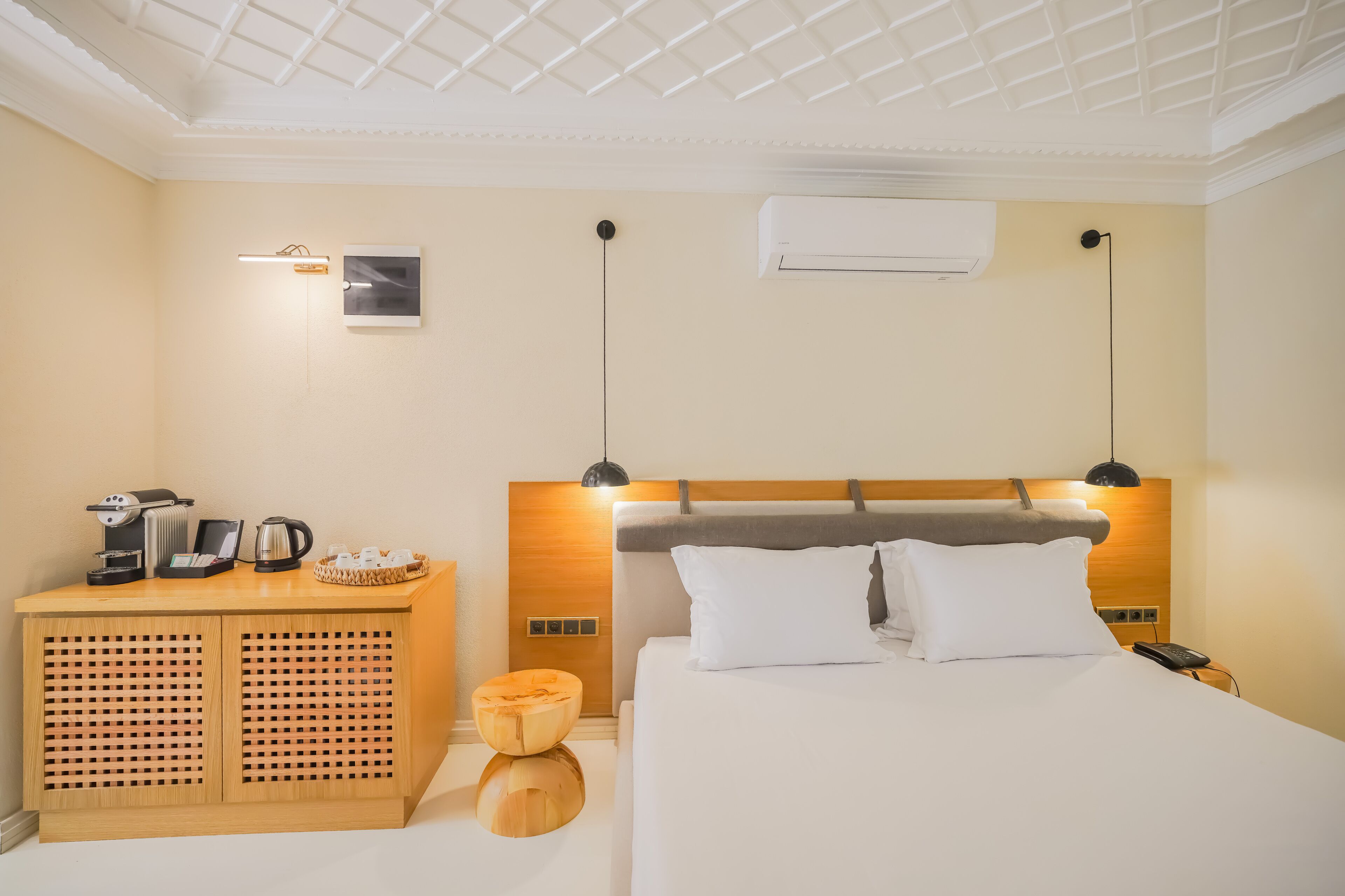 A hotel room with a wooden headboard, a bedside table with a coffee maker, a lamp, and a small wooden stool.