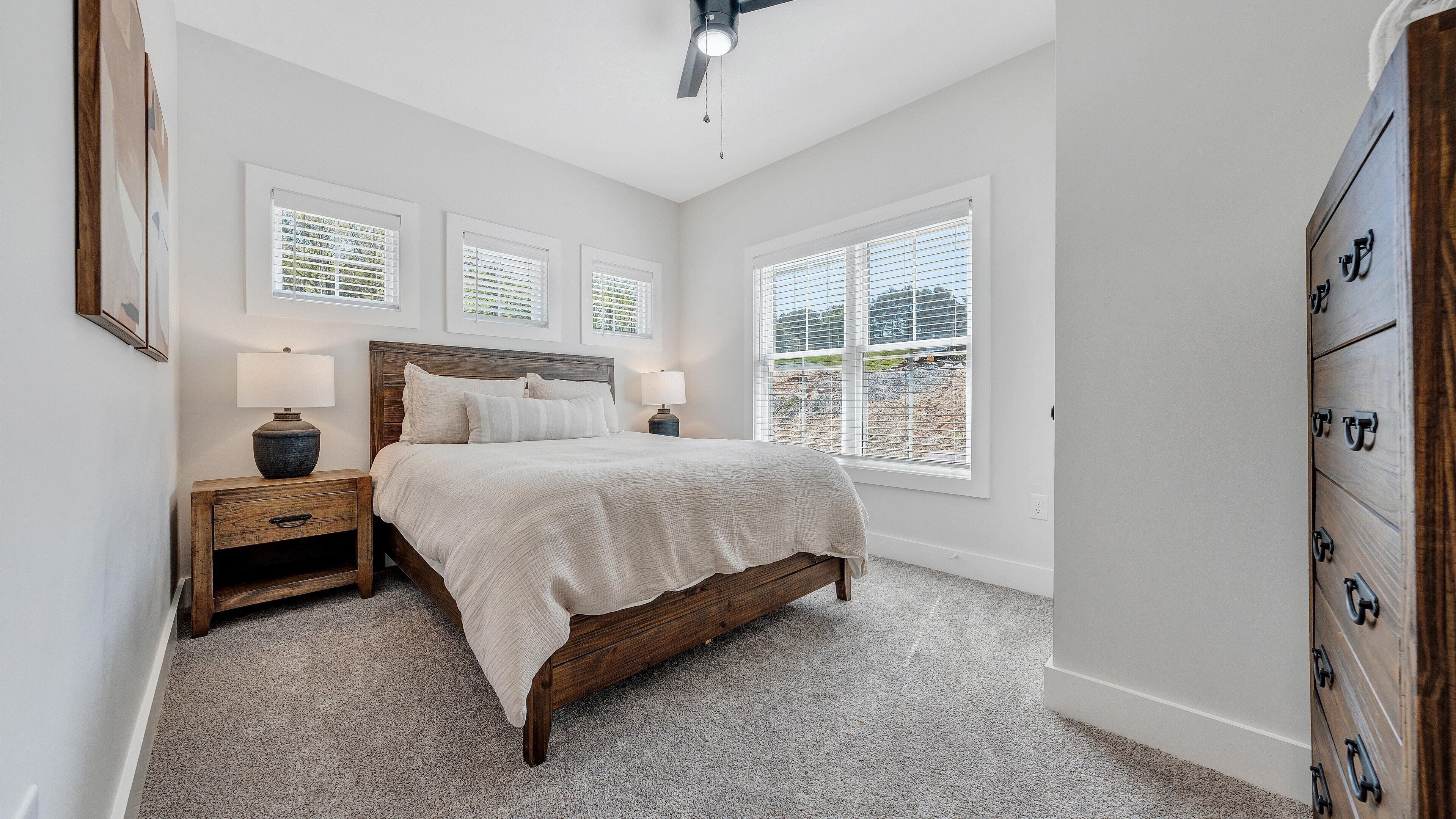 A bedroom with a large bed, two bedside tables, a dresser, and three windows with blinds.