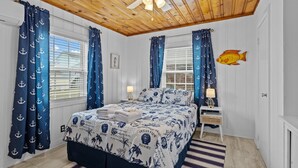 A bedroom with a wooden ceiling, a bed with a blue and white ocean-themed bedspread, and matching curtains. - The Saltbox-Home in Atlantic Beach (Atlantic Beach)