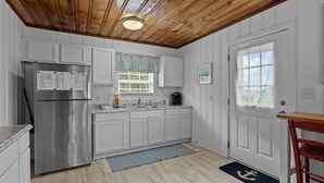 A kitchen with white cabinets, a stainless steel refrigerator, a window with a view, and a wooden ceiling. - The Saltbox-Home in Atlantic Beach (Atlantic Beach)
