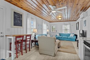 A cozy living room with a blue sofa, a wicker chair, a wooden ceiling, and a chalkboard sign. - The Saltbox-Home in Atlantic Beach (Atlantic Beach)