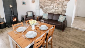 A dining area with a wooden table set for four, a stone wall, a fireplace, and a leather sofa. - Dairy Cottage (Whitland)