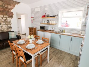 A modern kitchen with a wooden dining table set for four, light blue cabinets, a brick wall, and a window with a view of a red car. - Dairy Cottage (Whitland)