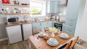 A modern kitchen with a wooden table set for dining, a dishwasher, a microwave, and light blue cabinets. - Dairy Cottage (Whitland)