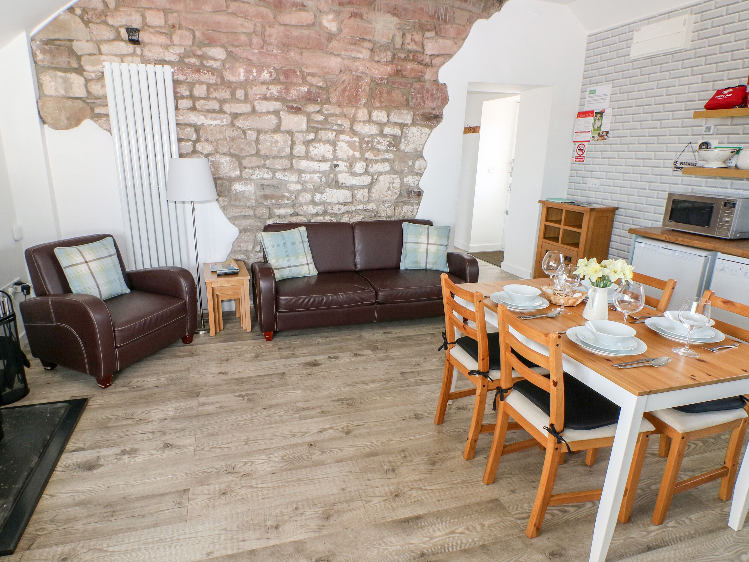 A cozy living room with a brown leather sofa, a wooden dining table set for four, and a kitchen area with a microwave and a refrigerator.