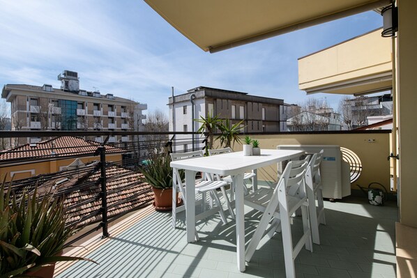 A balcony with a white table and chairs, potted plants, and a view of surrounding buildings.