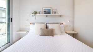 Slaapkamer met een groot bed, een raam, een nachtkastje met een lamp en een plank met foto’s en een plant.
