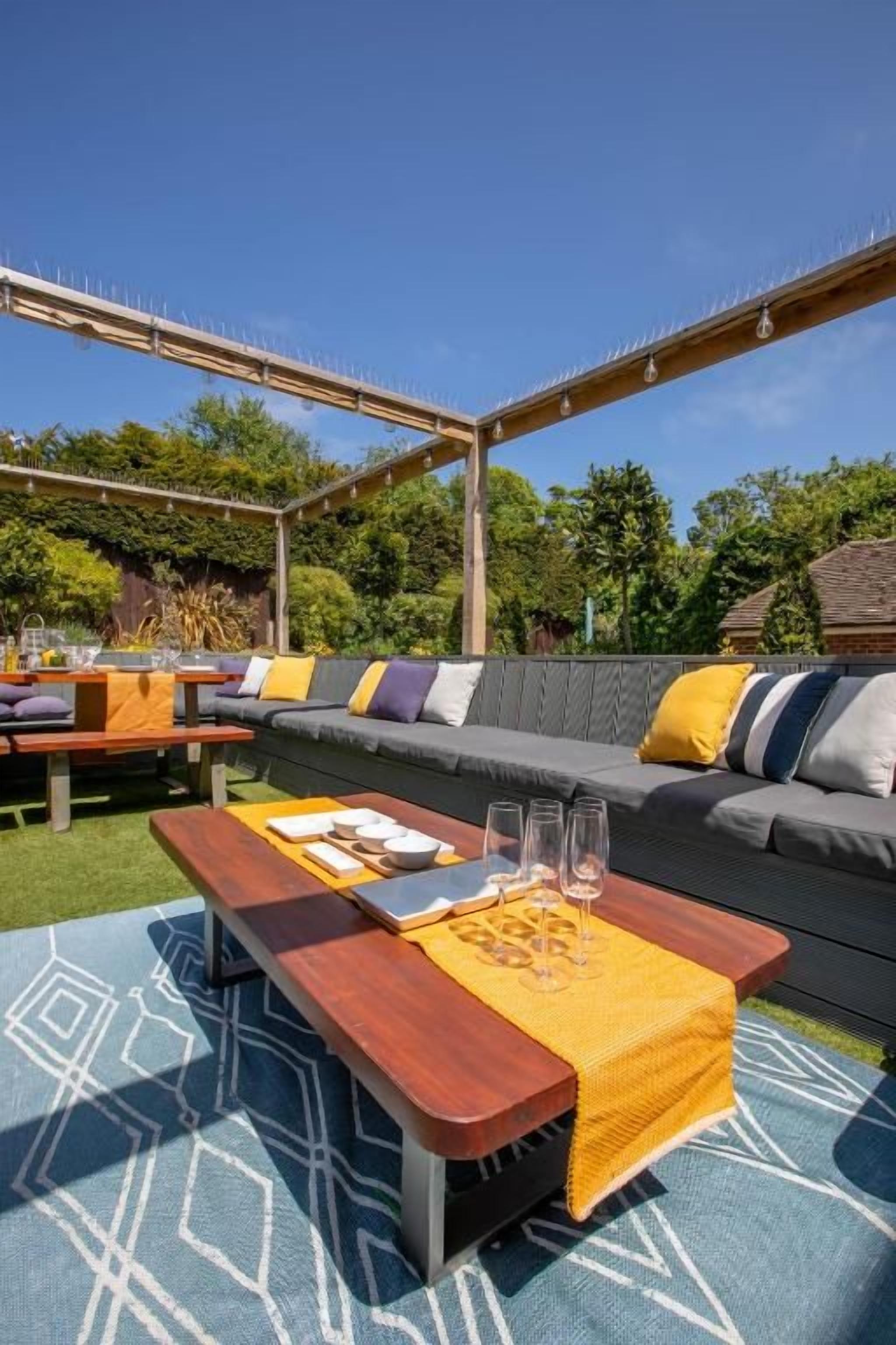 An outdoor seating area with a wooden pergola, a sofa with cushions, and a table with glasses and a menu.