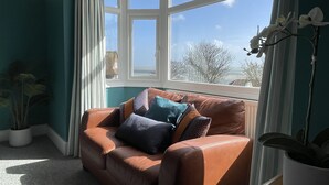 A room with a brown leather sofa, a bay window, and a view of trees and buildings outside. - Winterstoke View, Family and dog Friendly Beach Retreat (Ramsgate)