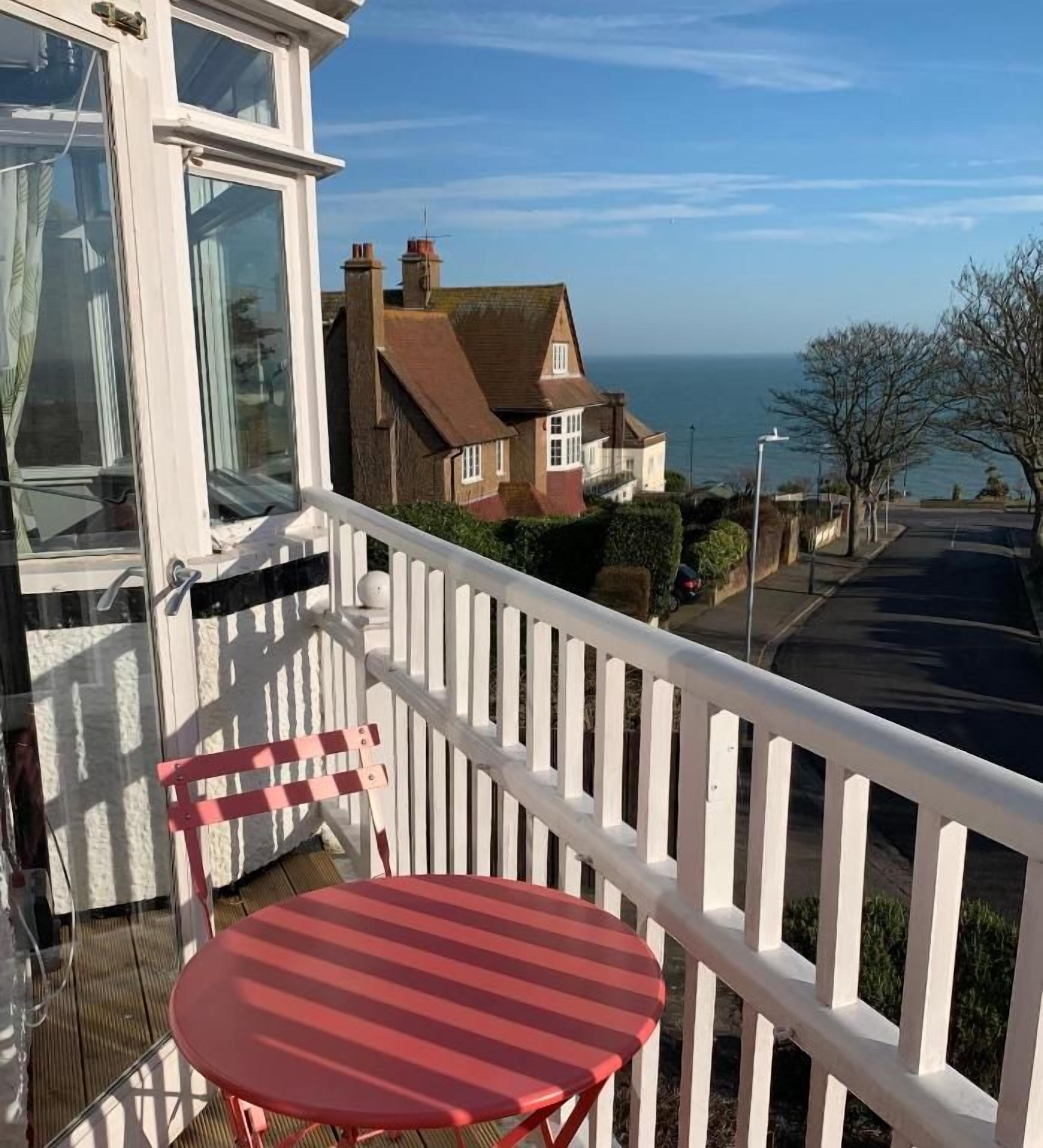 A balcony with a red chair and a white railing overlooking a residential street and the sea.