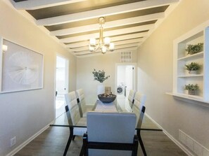 A dining area with a glass table, cushioned chairs, a chandelier, and a potted plant.