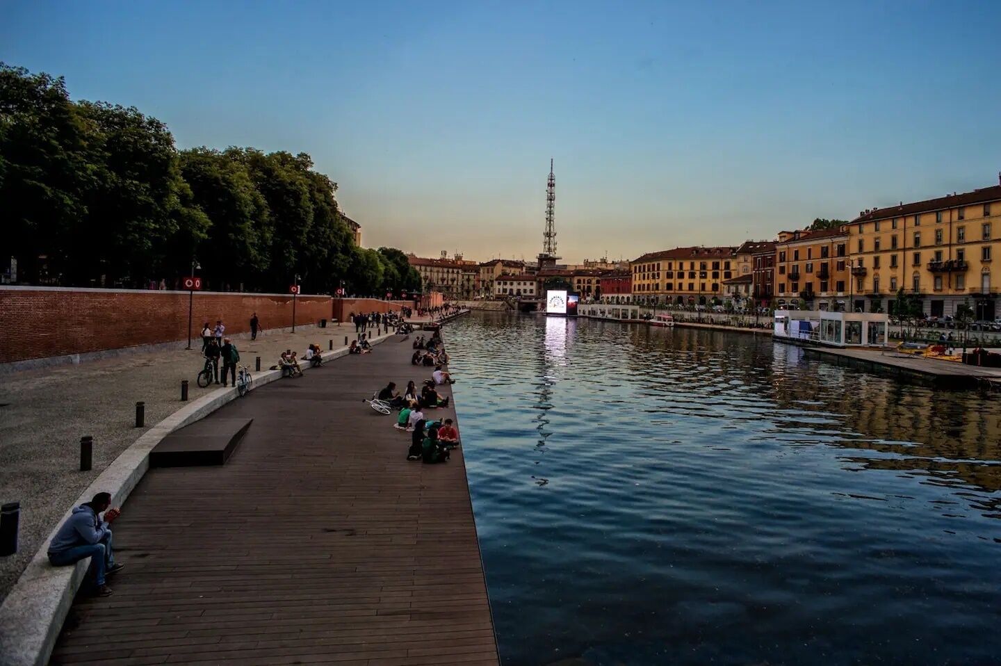 Una passeggiata lungo il fiume con una passerella in legno, persone sedute e che passeggiano, e edifici lungo l'argine.