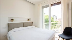 Ein Schlafzimmer mit einem großen Bett, einem Fenster mit Blick auf Gebäude und einem Sessel.