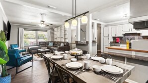 Een moderne keuken met witte kastjes, een eetgedeelte met een houten tafel voor vier personen, en een woonkamer met een blauwe bank en een plafondventilator.