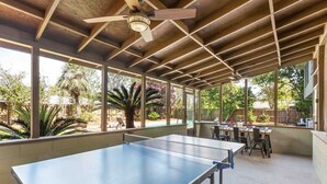 Een binnentafeltennistafel met plafondventilator, omringd door tafels en stoelen, onder een houten dak met grote ramen.
