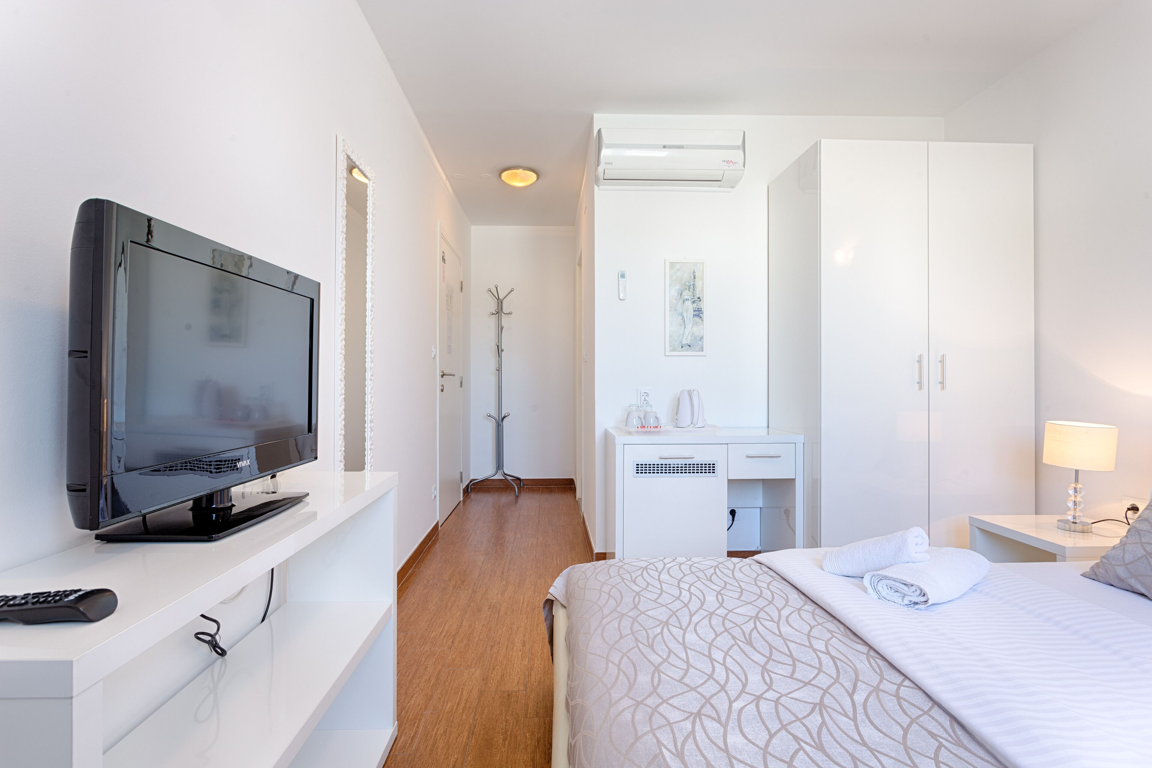 A hotel room with a flat-screen TV, a desk with a chair, a bed with a patterned bedspread, and a white wardrobe.