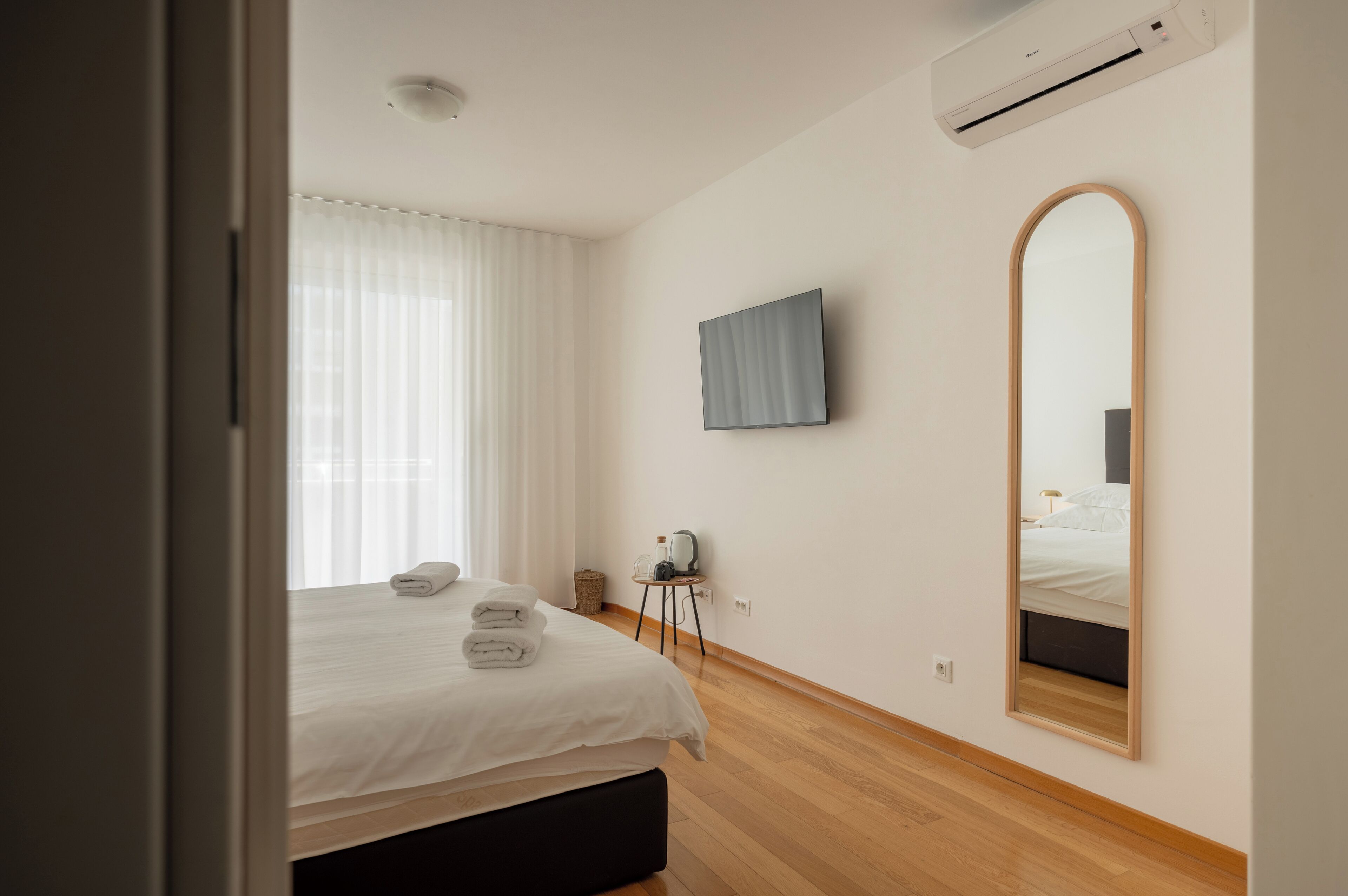 Una habitación de hotel moderna con una cama, un televisor de pantalla plana, un espejo grande y una mesita de noche con lámpara.