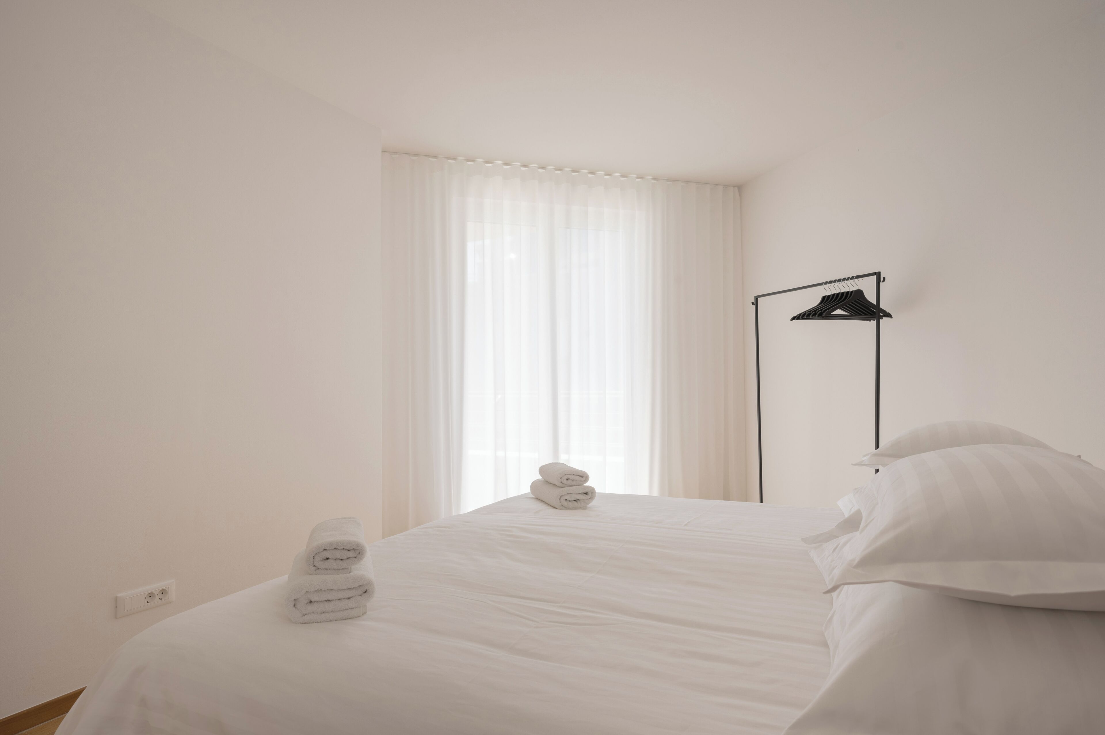 A neatly made bed with white linens and rolled towels, a black hanger on a stand, and a window with sheer curtains.